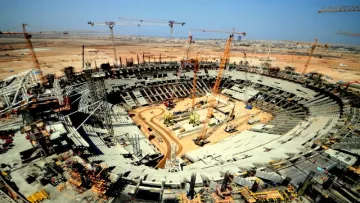 L'image montre un immense chantier de construction d'un stade. On peut voir une grande arène en forme de cercle, avec des gradins en cours d'assemblage. De nombreuses grues sont présentes, surélevant des matériaux de construction, et divers engins lourds sont dispersés sur le site. À l'horizon, on aperçoit des bâtiments et un ciel dégagé, indiquant une propreté et un bon déroulement des travaux. L'ensemble de la scène illustre l'activité dynamique et l'envergure d'un projet architectural ambitieux.