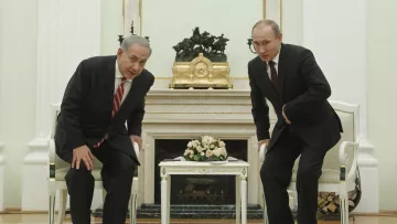 L'image montre deux hommes en costume, l'un avec une cravate rayée et l'autre avec une cravate sombre, se tenant debout devant une table basse dans une pièce élégante. Ils semblent se pencher légèrement en avant, probablement lors d'une conversation. En arrière-plan, on peut voir des éléments de décoration intérieure, dont une grande horloge et un bouquet de fleurs sur la table. Les murs sont peints dans des tons clairs et l'ensemble donne une impression de formalité.