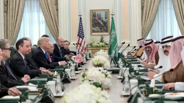 L'image montre une réunion formelle dans une grande salle avec une longue table. D'un côté, plusieurs représentants américains, dont on peut distinguer deux hommes en costume, ainsi qu'une personne qui semble être le président. En face d'eux, des dignitaires saoudiens, également en costume traditionnel. La salle est ornée de drapeaux américain et saoudien, et des arrangements floraux décoraient la table. L'atmosphère semble sérieuse et collaborative.