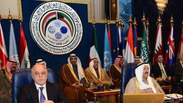 The image depicts a formal conference setting, featuring several dignitaries sitting in a room with flags from various nations displayed prominently in the background. The attendees are dressed in traditional attire, indicating a Middle Eastern cultural context. A large emblem or logo is visible above them, likely representing the conference's purpose, which involves international cooperation or reconstruction efforts. Among the seated figures, one appears to be a prominent leader or official, positioned at the forefront of the image. The overall atmosphere suggests a significant meeting aimed at addressing important regional or global issues.