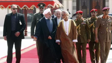 L'image montre un groupe de personnes marchant sur un tapis rouge. Au centre, deux hommes en discussion, l'un portant une robe noire et l'autre une robe traditionnelle d'Oman. Ils sont entourés de plusieurs membres de la sécurité et d'officiers en uniforme. Le cadre est formel, avec un décor architectural en arrière-plan qui suggère un événement diplomatique ou officiel.