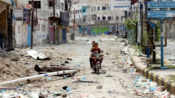 L'image montre une scène de rue désolée et abandonnée, probablement dans une zone urbaine touchée par des conflits ou des désastres. Le sol est recouvert de débris et de détritus, tandis que les bâtiments autour semblent endommagés. Au centre de l'image, deux personnes sont assises sur une moto, traversant cette rue dévastée. Les enseignes et les murs portent des marques de détérioration, reflétant un environnement difficile et chaotique.