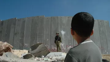 در این تصویر، پسر بچه‌ای به سمت یک دیوار بلند و بتنی نگاه می‌کند. دیوار دارای آثار graffiti و علامت‌های مختلفی است. در مقابل او، یک نظامی ایستاده است که لباس نظامی به تن دارد و به نظر می‌رسد در حال نظارت است. محیط اطراف شامل سنگ‌ها و زباله‌های پراکنده است و آسمان آبی و روشنی بالای سر آنها دیده می‌شود. احساس تصویر می‌تواند تنش و جدایی را به خوبی نشان دهد.