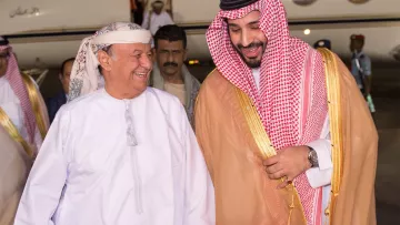 The image shows two men smiling and engaged in a friendly conversation as they walk towards a private jet. One man is dressed in traditional Arab attire, including a white thobe and a headscarf, while the other is wearing a similar traditional outfit, accented with a golden cloak and a checkered headscarf. They appear to be happy and are holding hands, suggesting a warm greeting. In the background, there are several individuals, likely staff or security, standing by the jet. The setting seems to be an airport or a private aviation terminal.