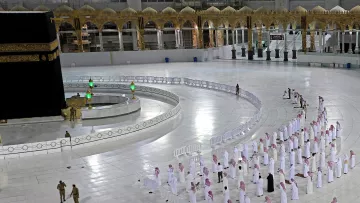 La imagen muestra el interior de la Gran Mezquita de La Meca, donde se encuentra la Kaaba. Se puede ver un gran espacio blanco con filas de personas en posición de oración. Están vistiendo tradicionalmente con túnicas blancas y pañuelos en la cabeza. En el fondo, se aprecia la Kaaba con su cubierta negra y algunos elementos decorativos. También hay barreras que delimitan el espacio y un par de agentes de seguridad en el área. La atmósfera es de calma y devoción.