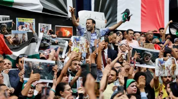 L'image montre une foule rassemblée lors d'une manifestation, avec des personnes brandissant des pancartes et des photos. Au centre, un homme lève les bras en signe de victoire ou de protestation. La foule exprime visiblement des émotions fortes, que ce soit la colère ou la solidarité. Des drapeaux sont également présents dans le décor, ajoutant à l'intensité de la scène. On ressent une atmosphère d'engagement et de mobilisation collective.