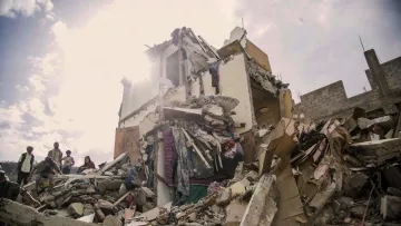 The image depicts a collapsed building, showing significant structural damage with debris scattered around. The upper portions of the structure are crumbled, and various materials like concrete, wood, and personal belongings are visible. In the foreground, there are a few individuals, likely responders or onlookers, inspecting the site. The scene is illuminated by sunlight breaking through the clouds, which adds a stark contrast to the devastation shown. The overall mood conveys a sense of disaster and the aftermath of a catastrophic event.