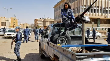 L'image montre une scène urbaine impliquant des forces de sécurité. On peut y voir plusieurs policiers en uniforme, certains armés, se déplaçant autour de véhicules. Un homme est assis sur le plateau d'une camionnette, près d'une arme lourde montée sur le véhicule. L'environnement semble être une ville moderne, avec des bâtiments en arrière-plan. L'ambiance générale de l'image suggère une opération de sécurité ou une situation de contrôle.