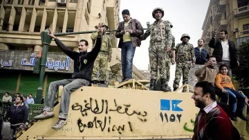 The image depicts a group of people, including soldiers, gathered on top of a military vehicle in an urban setting. Some individuals are gesturing and expressing emotions, while others appear to be observing or interacting with the crowd below. The environment is bustling, with buildings visible in the background, indicating a scene of protest or public demonstration. The atmosphere suggests a moment of tension or significant political activity.