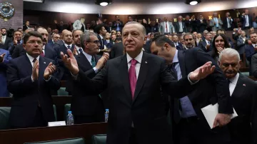 L'image montre un homme en costume, les bras écartés, se tenant au centre d'une assemblée constituée de nombreuses personnes applaudissant autour de lui. Il semble apprécier l'attention du public, dans un environnement de type parlementaire, où les membres sont assis sur des bancs et affichent des expressions variées. L'atmosphère est dynamique, suggérant un moment de célébration ou de discours important.