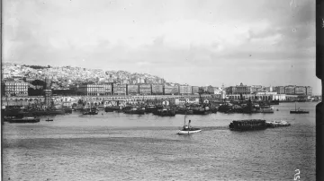 L'image représente un port où l'on peut voir plusieurs bateaux amarrés. Au fond, une ville s'étend, avec des bâtiments alignés sur la côte. Le ciel est nuageux, et l'eau calme reflète la scène. Les collines derrière la ville sont habitées, ajoutant à la densité urbaine de l'endroit. L'ambiance générale semble nostalgique, probablement en raison du style photographique ancien.