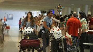 L'image montre une scène dans un aéroport, avec des voyageurs. À l'avant, un homme pousse une chariot de bagages et une femme derrière lui, également avec un chariot. Ils semblent attendent ou se déplacent parmi d'autres passagers. En arrière-plan, on aperçoit une longue file de personnes, probablement en attente de faire enregistrer leurs bagages ou de passer la sécurité. L'ambiance semble animée, typique des aéroports.