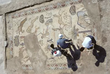 L'image montre une vue aérienne d'un sol en mosaïque impressionnant, découvert lors de fouilles archéologiques. Cette mosaïque représente une variété d'animaux, tels que des lions et des cerfs, ainsi que des motifs floraux et géométriques. On peut voir deux personnes travaillant à proximité, probablement en train d'examiner ou de restaurer la mosaïque. La terre autour de la mosaïque est dénudée, suggérant un site archéologique en plein exploration. Les ombres des personnes ajoutent une dimension intéressante à la scène.