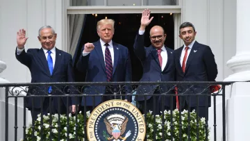 L'image montre quatre hommes debout sur un balcon de la Maison Blanche, levant la main en signe de salutation. Ils semblent être en train de célébrer un moment important, probablement lié à des événements diplomatiques. L'un des hommes porte un costume sombre avec une cravate rouge, tandis que les autres affichent également des tenues formelles. En arrière-plan, on peut voir un environnement architectural caractéristique de la Maison Blanche, et un grand sceau du Président des États-Unis est en évidence sur la balustrade.