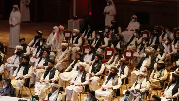 La imagen muestra a un grupo de personas sentadas en un auditorio. Están vestidos con prendas tradicionales, como túnicas y turbantes. La mayoría lleva mascarillas, probablemente debido a precauciones de salud. El ambiente parece ser formal y puede estar relacionado con una reunión o conferencia. Al fondo, se pueden ver más asistentes de pie. La disposición de los asientos sugiere un evento importante, probablemente político o social.