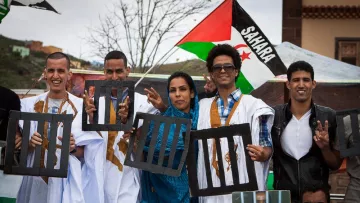 L'image montre un groupe de personnes posant ensemble lors d'une manifestation. Ils portent des vêtements traditionnels, notamment des robes blanches. Certains affichent des signes pacifiques avec leurs mains, tandis que d'autres tiennent des panneaux représentant des barreaux de prison. En arrière-plan, on peut voir un drapeau aux couleurs du Sahara. L'atmosphère semble engagée et solidaire.