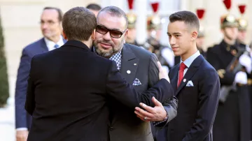 The image features a formal scene where two men are embracing, likely in a greeting or farewell. One man is wearing glasses and a suit, and he appears to be the primary focus of the hug. The other man is dressed in a dark coat and seems to be extending his arms in a friendly gesture. In the background, there are other individuals in formal attire, including guards. There's a young man standing beside the two men, dressed in a suit with a tie, looking on. The setting seems to be a ceremonial occasion, possibly outside a governmental building.