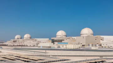 L'immagine mostra una centrale nucleare moderna, caratterizzata da tre imponenti cupole bianche che emergono dal complesso. La struttura presenta un design industriale, con vari edifici di colore chiaro che si estendono in un'area ampia e ben organizzata. Il cielo è sereno e blu, suggerendo una bella giornata. In primo piano, si può notare un'area ben curata e delle strade di accesso, tipiche delle installazioni di questo tipo.