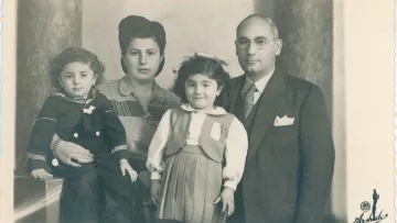 Cette image en noir et blanc montre une famille posant ensemble. À gauche, un jeune garçon portant une tenue de marin est assis. À côté de lui, une femme au style classique avec des cheveux coiffés en pompadour se tient. En face d'elle, une petite fille avec un sourire doux porte une robe avec un gilet. À droite, un homme en costume, portant des lunettes, regarde la caméra. L'arrière-plan est sobre, ajoutant une touche nostalgique à cette photo de famille.