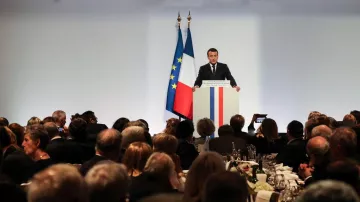 L'image montre un événement formel où une personne, probablement un homme politique, se tient devant un podium. Il parle à un public assis à des tables bien dressées, avec des couverts et des verres. En arrière-plan, on peut voir des drapeaux tricolores, bleu, blanc et rouge, représentant la France. L'atmosphère semble être celle d'un dîner officiel ou d'une réception. Les invités sont attentifs et nombreux, reflétant un moment de discours ou de présentation dans un cadre élégant.