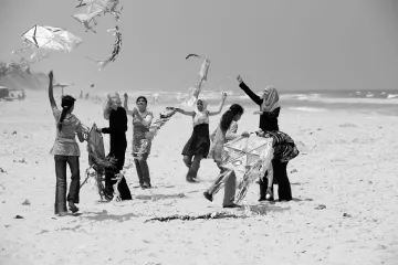 L'image montre un groupe de femmes sur une plage, s'amusant à faire voler des cerfs-volants. Certaines d'entre elles sont en train de tenir ou de manipuler des cerfs-volants, tandis que d'autres semblent rire et profiter du moment. L'environnement est ensoleillé, avec des vagues en arrière-plan. La scène dégage une atmosphère joyeuse et festive, mettant en avant des interactions sociales et des activités de plein air.