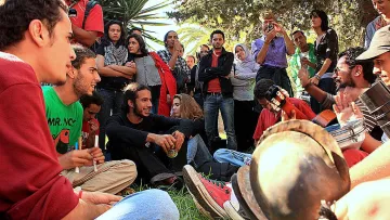 L'image montre un groupe de personnes rassemblées dans un espace extérieur, probablement un parc. Certains d'entre eux sont assis sur le sol, tandis que d'autres se tiennent debout ou sont légèrement en retrait. La scène semble conviviale et décontractée, avec des gens qui discutent, rient ou écoutent de la musique. Au centre, on aperçoit des instruments de musique, suggérant qu'il y a une performance musicale en cours. Les personnes présentes semblent diverses, avec des styles vestimentaires variés, ce qui indique une atmosphère informelle et communautaire.