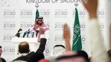 L'image montre une conférence de presse organisée en Arabie Saoudite. Un homme, vêtu d'une robe traditionnelle saoudienne et d'un headdress, se tient devant un podium avec plusieurs microphones. En arrière-plan, on peut voir des drapeaux saoudiens ainsi qu'un panneau affichant des informations sur la Vision 2030 de l'Arabie Saoudite. Plusieurs personnes sont présentes, levant les mains, probablement pour poser des questions. L'atmosphère semble sérieuse, centrée sur des discussions importantes liées à l'avenir du pays.