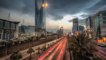 L'image montre une vue urbaine d'une grande ville, probablement Riyad, avec des gratte-ciel modernes qui se dressent contre un ciel nuageux. On peut voir une route animée avec des phares de voitures qui créent des traînées lumineuses, suggérant une circulation intense. Des palmiers sont visibles le long de la route, ajoutant une touche naturelle à ce paysage urbain. La scène est éclairée par des lampadaires, donnant une atmosphère à la fois dynamique et contemporaine.