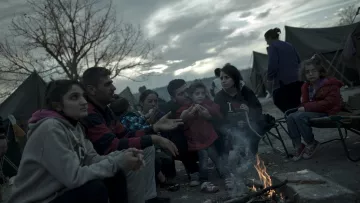 L'image montre un groupe de personnes assises autour d'un feu de camp, probablement dans un camp de réfugiés ou un site de fortune. On peut voir des adultes et des enfants, certains vêtus de manteaux pour se protéger du froid. L'ambiance semble sombre en raison des nuages menaçants dans le ciel. Les visages des personnes expriment une variété d'émotions, allant de la tristesse à l'espoir. En arrière-plan, on aperçoit des tentes qui indiquent que ce groupe vit dans des conditions temporaires.