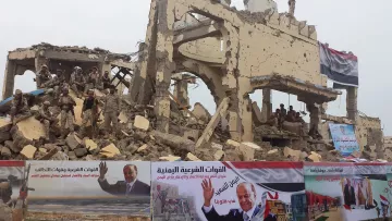 L'image montre un bâtiment en ruines, probablement à la suite d'un conflit ou d'une destruction. Des soldats sont visibles parmi les débris, ce qui indique une présence militaire dans la zone. Au premier plan, des affiches sont accrochées, probablement en soutien à un leader ou un mouvement, avec du texte en arabe. L'ensemble évoque une atmosphère de résilience et de reconstruction au milieu des dégâts.