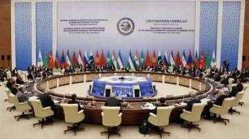 The image depicts a large conference room set up for a high-level meeting, with a circular table at the center surrounded by delegates. Various flags representing different countries are displayed around the room, indicating a multinational gathering. Attendees, likely officials or representatives, are seated and engaged in discussion. The backdrop features stylized graphics and text, suggesting it is related to a significant international conference, possibly focusing on cooperation among member states. The atmosphere appears formal and collaborative.