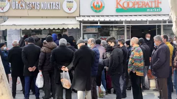 L'image montre une foule de personnes faisant la queue devant un magasin, probablement un marché ou un point de vente. Les gens semblent attendre pour entrer ou acheter des produits. Ils portent des vêtements d'hiver, et certains tiennent des sacs. L'atmosphère semble calme, mais il y a une certaine impatience dans la file d'attente. Au-dessus de l'entrée du magasin, on peut voir des panneaux indiquant le nom et la fonction de l'établissement.