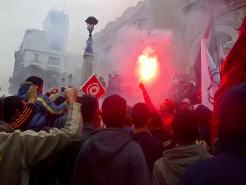 L'image montre une foule rassemblée dans une manifestation. Des personnes tiennent des drapeaux, dont un drapeau tunisien, et certains agitent des fumigènes qui dégagent de la fumée rouge. L'ambiance semble énergique et engagée, avec des bâtiments en arrière-plan. La foule paraît déterminée et unie autour de leur cause.
