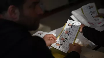 L'image montre une personne tenant un bulletin de vote. En arrière-plan, on peut voir d'autres bulletins similaires. Les bulletins présentent des portraits de candidats avec leurs noms et éventuellement des informations liées à l'élection. L'ambiance semble être celle d'un bureau de vote. La lumière est naturelle et le cadre semble simple, typique d'une situation électorale.