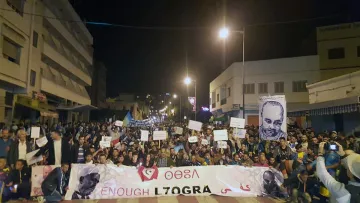 L'image montre une grande manifestation nocturne dans une rue animée. Beaucoup de personnes sont rassemblées, tenant des pancartes et banderoles. On peut voir des portraits de plusieurs individus exposés parmi les manifestants. L'atmosphère semble énergique et engagée, avec des participants qui expriment leurs revendications. Des lumières artificielles illuminent la scène, créant une ambiance dynamique.