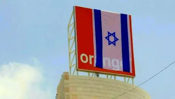 L'image montre un panneau affichant le logo de la marque "Orange", sur lequel est drapé un grand drapeau israélien. Le fond semble être un ciel clair, et le drapeau présente des bandes horizontales en bleu et blanc avec l'étoile de David au centre. Le tout donne une impression d'affichage public dans un environnement urbain.