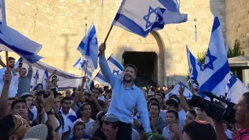 L'image montre une foule rassemblée devant une grande porte en pierre, probablement située dans une ville historique. Les participants tiennent des drapeaux bleu et blanc, représentant l'État d'Israël. Un homme se tient au centre, soulevant un drapeau avec un sourire, tandis que d'autres l'entourent, festoyant. L'atmosphère semble festive et joyeuse, suggérant une célébration ou un événement important.