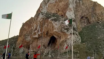 La imagen muestra un paisaje montañoso con una gran roca o acantilado en el centro, que tiene una cueva visible en su base. A su alrededor, hay varias banderas ondeando, entre las cuales algunas son de Argelia, ya que tienen un diseño distintivo. En la parte inferior de la imagen, se pueden ver algunas personas caminando y disfrutando del lugar. El cielo tiene un tono claro, posiblemente al atardecer. La escena refleja un ambiente natural y un sitio de interés cultural o histórico.