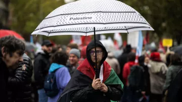 The image depicts a gathering, likely a protest or demonstration, where a person stands under a black-and-white umbrella labeled "Palestine." This individual is draped in a Palestinian flag and wears glasses, displaying a serious expression. Surrounding them are other participants, some holding flags and signs, with a backdrop of trees and a cloudy sky, suggesting an outdoor setting and possibly inclement weather. The scene captures a moment of solidarity and advocacy related to Palestinian issues.