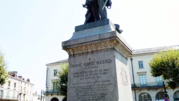 L'image représente une statue érigée à la mémoire de Thomas Robert Bugeaud. La statue est placée sur un socle en pierre portant une inscription détaillant ses accomplissements. Bugeaud est présenté en tenue militaire, tenant son épée. En arrière-plan, on peut voir une rue avec des arbres et des bâtiments, indiquant probablement un espace public. La lumière est claire, suggérant une journée ensoleillée.