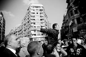 The image depicts a scene of a protest or demonstration. In the foreground, a young man is animatedly speaking or shouting, with his arms outstretched, indicating passion or urgency. Surrounding him are several people, some looking up at him with expressions of engagement or concern. The background features tall buildings, hinting at an urban environment. The photograph is in black and white, which emphasizes the intensity and emotions of the moment. The overall atmosphere conveys a sense of solidarity and activism.