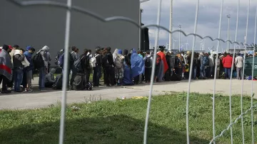 L'image montre une file de personnes faisant la queue près d'un bâtiment, probablement en attendant de recevoir de l'aide ou des services. Les personnes sont vêtues de diverses manières, avec certains portant des couvertures. On peut voir une clôture en fil de fer qui sépare le groupe du reste de l'environnement. En arrière-plan, on aperçoit des installations modernes et des éoliennes, suggérant une zone urbaine ou industrielle. Le ciel est nuageux, ce qui pourrait indiquer une atmosphère morose.