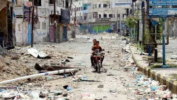 The image depicts a deserted street scene that appears to be in disrepair and heavily littered. There is debris scattered across the ground, suggesting neglect or the aftermath of a conflict. A couple of children can be seen riding a motorcycle down the street, which seems to be a common mode of transportation in the area. Buildings along the street show signs of damage, with faded signs and graffiti on the walls. Overall, the atmosphere conveys a sense of abandonment and deterioration.