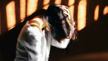 L'image montre une femme assise, penchée en avant et se tenant la tête avec ses mains. Sa posture suggère un état de réflexion ou de malaise. La lumière du soleil filtre à travers des barreaux, créant des ombres graphiques sur elle et sur le mur derrière. Elle porte un vêtement doux et pelucheux, ce qui contraste avec l'intensité de la lumière. L'ambiance générale de l'image évoque une certaine introspection ou une atmosphère mélancolique.