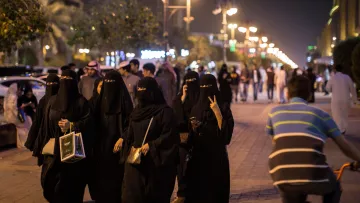 L'image montre un groupe de femmes vêtues de tenues noires traditionnelles marchant ensemble sur un trottoir. Certaines d'entre elles portent des masques qui dissimulent leur visage, tandis que d'autres affichent des gestes amicaux. En arrière-plan, on voit une rue animée avec des passants et des lumières, évoquant une ambiance nocturne. Un homme à vélo passe à proximité, ajoutant à la dynamique de la scène.