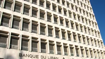 L'image montre un bâtiment moderne qui abrite la Banque du Liban. La façade est caractérisée par un design géométrique, avec de nombreuses fenêtres disposées en grille, donnant une impression de symétrie. On peut également voir le nom de la banque inscrit en arabe et en français, ainsi qu'un symbole représentant un cèdre, un arbre emblématique du Liban. L'architecture est sobre et imposante, typique des institutions financières. Le ciel est clair, suggérant une journée ensoleillée.