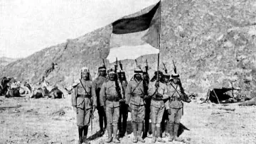 L'image représente un groupe de soldats posant devant un drapeau. Ils sont vêtus de uniforme militaire, portant des capes et des casques. En arrière-plan, on aperçoit un paysage montagneux et quelques tentes, suggérant un camp militaire. Les soldats, tenant leurs fusils, semblent être en position de parade ou de cérémonie. L'atmosphère de la photo évoque une période historique de conflit ou de guerre.