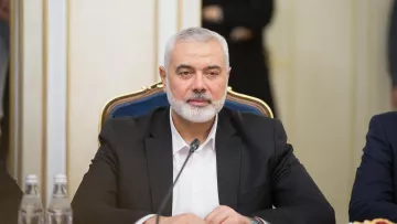 در تصویر، مردی با ریش خاکستری و موهای کوتاه دیده می‌شود که در حال نشستن در یک جلسه یا کنفرانس است. او ظاهری جدی دارد و یک کت تیره به تن دارد. بر روی میز مقابل او بطری‌های آب و تعدادی خوراکی دیده می‌شود. فضای اطراف به نظر رسمی و مرتبط با مذاکرات یا بحث‌های مهم می‌رسد.