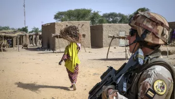 L'image montre un soldat armé en position de surveillance dans un environnement désertique. Il est vêtu d'un uniforme militaire et porte un casque. À l'arrière-plan, on aperçoit une femme portant un fardeau, sans doute des branches, qui s'éloigne pour se rendre à un village constitué de constructions en terre. Le terrain est sablonneux, et quelques arbres épars se trouvent aux alentours. Cette scène évoque un contexte de sécurité dans une région rurale.