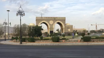 Un grand portail avec arches, entouré de verdure et de lampadaires, sous un ciel nuageux.